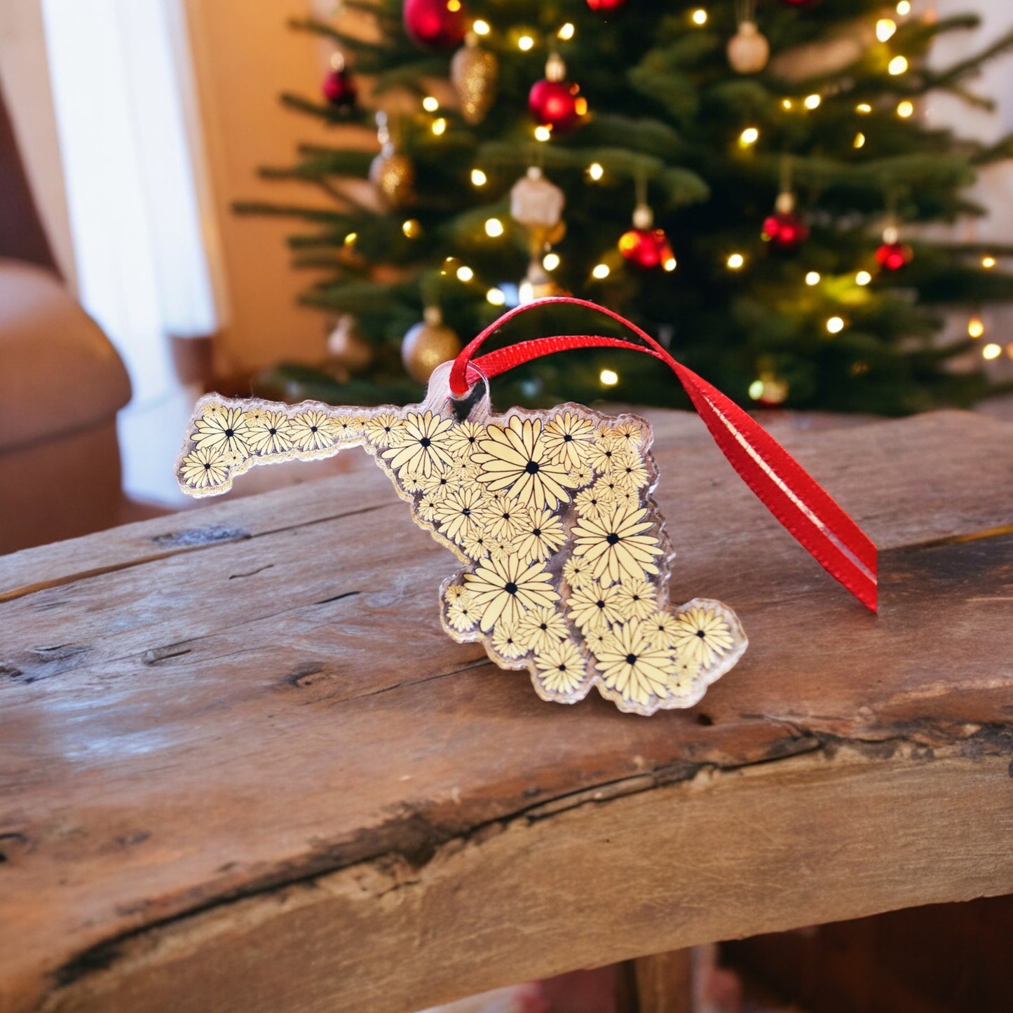 acrylic Christmas ornament shaped like the state of Maryland covered in black eyed susans with a red ribbon holder. ornament is propped up on a wood table in front of a decorated Christmas tree